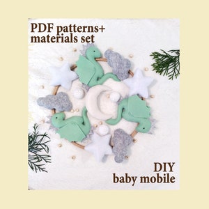 May include: A DIY baby mobile featuring felt dinosaurs, stars, clouds, and a crescent moon in shades of green, white, and gray. The mobile is suspended on a wooden ring. Text on the image reads "PDF patterns+ materials set" and "DIY baby mobile."