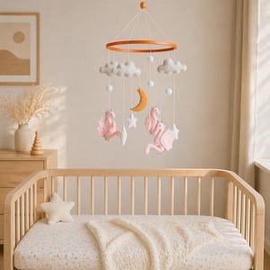 May include: A baby crib with a wooden frame and a cream-colored fitted sheet and blanket. Above the crib hangs a mobile with pink dragons, white clouds, stars, and a wooden crescent moon. The mobile is suspended from a wooden ring.