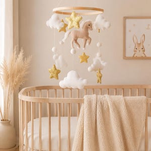 May include: A baby crib with a beige blanket and a hanging mobile. The mobile features a wooden ring, a plush horse, white clouds, gold stars, and white spheres. A framed bunny art piece is visible.