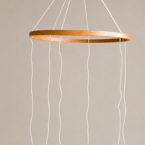 May include: A hanging mobile frame consisting of a circular wooden hoop suspended by white strings that meet at a central wooden bead. Multiple white strings hang vertically from the hoop, creating a structure for displaying decorative items.