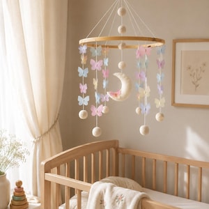 May include: A baby mobile with a wooden hoop, featuring pastel-colored butterfly cutouts, felt balls, and a crescent moon. The mobile hangs above a wooden crib with a floral-embroidered blanket. The room has a neutral color palette.