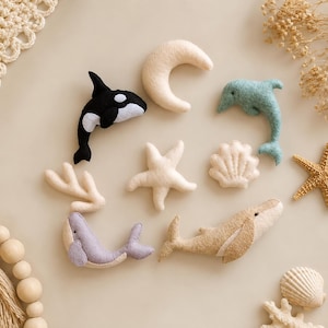 Peut inclure: Une collection d'ornements en feutrine sur le thème de l'océan comprenant un orque noir et blanc, un dauphin bleu clair, une baleine beige, une baleine lavande, une lune crème, une étoile de mer, un coquillage et une branche de corail.
