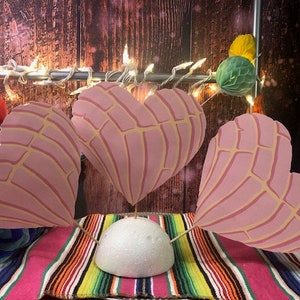 May include: Three pink heart-shaped decorations with a textured pattern, resembling a traditional Mexican sweet bread, are displayed on a colorful striped blanket. The decorations are arranged in a triangular shape, with the base of the triangle resting on the blanket. The decorations are held up by wooden skewers, and the tips of the skewers are inserted into a white styrofoam base.