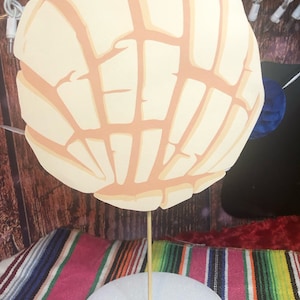 May include: A decorative, oversized concha pastry cutout, featuring a light beige base with tan-colored lines, mounted on a white foam base with a wooden dowel. The background includes a colorful striped textile and a wooden surface.