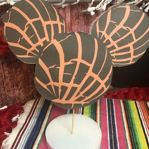 May include: Three-piece concha-shaped decorations in a Mickey Mouse head design. The conchas are dark gray with orange-tan lines, mounted on a white foam base with a wooden skewer. The background includes a colorful striped textile and fairy lights.