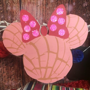 May include: A pink paper cutout resembling a Minnie Mouse head, with two concha-shaped ears and a red bow adorned with pink glitter circles. The background is a wooden surface with string lights.