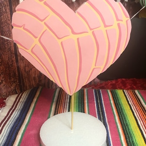 May include: A pink and yellow heart-shaped cake topper with a decorative design. The topper is on a white cake stand and a colorful striped tablecloth.