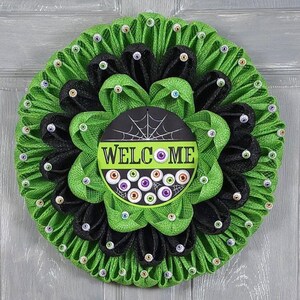 May include: A green and black Halloween wreath with a spiderweb and the word "WELCOME" on a black circle. The wreath is decorated with googly eyes.