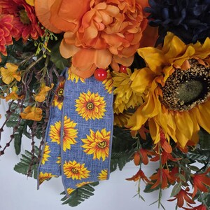 Puede incluir: Una corona decorativa con grandes peon&iacute;as naranjas, girasoles amarillos y flores azul oscuro. Una cinta azul con un patr&oacute;n de girasoles amarillos cuelga por el centro, acentuada por peque&ntilde;as bayas rojas y follaje verde en todo el arreglo.