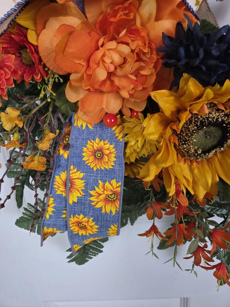 Puede incluir: Una corona decorativa con grandes peon&iacute;as naranjas, girasoles amarillos y flores azul oscuro. Una cinta azul con un patr&oacute;n de girasoles amarillos cuelga por el centro, acentuada por peque&ntilde;as bayas rojas y follaje verde en todo el arreglo.