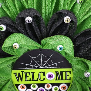 May include: A green and black Halloween wreath with a welcome sign and googly eyes. The sign has a spiderweb design and the word "WELCOME" in white letters.
