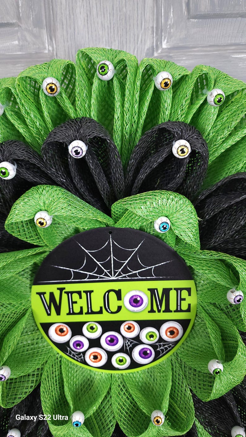 May include: A green and black Halloween wreath with a welcome sign and googly eyes. The sign has a spiderweb design and the word "WELCOME" in white letters.