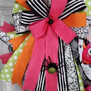 Puede incluir: Una corona de Halloween con un lazo rosa y una ara&ntilde;a brillante. La corona presenta cintas en patrones rosas, naranjas, verdes y blancos y negros, incluyendo lunares, rayas y telara&ntilde;as. Una decoraci&oacute;n festiva para la temporada de miedo.