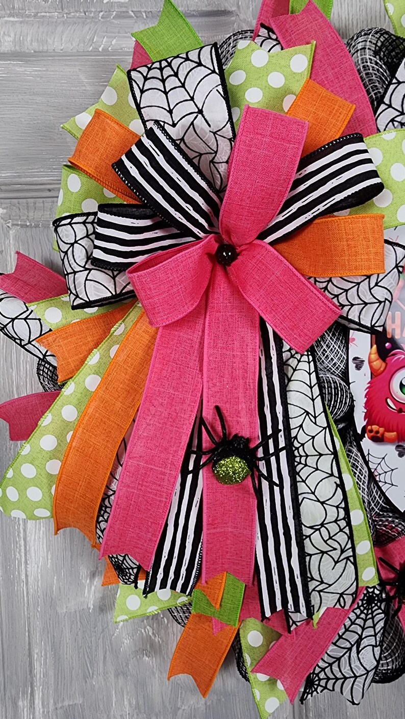 Puede incluir: Una corona de Halloween con un lazo rosa y una ara&ntilde;a brillante. La corona presenta cintas en patrones rosas, naranjas, verdes y blancos y negros, incluyendo lunares, rayas y telara&ntilde;as. Una decoraci&oacute;n festiva para la temporada de miedo.