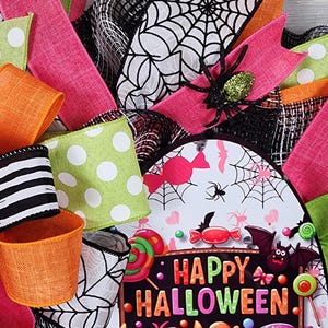 Puede incluir: Una corona de Halloween con cintas coloridas en rosa, verde y naranja. La corona presenta un letrero que dice "Happy Halloween" con monstruos de dibujos animados, murci&eacute;lagos y caramelos. Tambi&eacute;n se incluyen ara&ntilde;as y telara&ntilde;as decorativas.