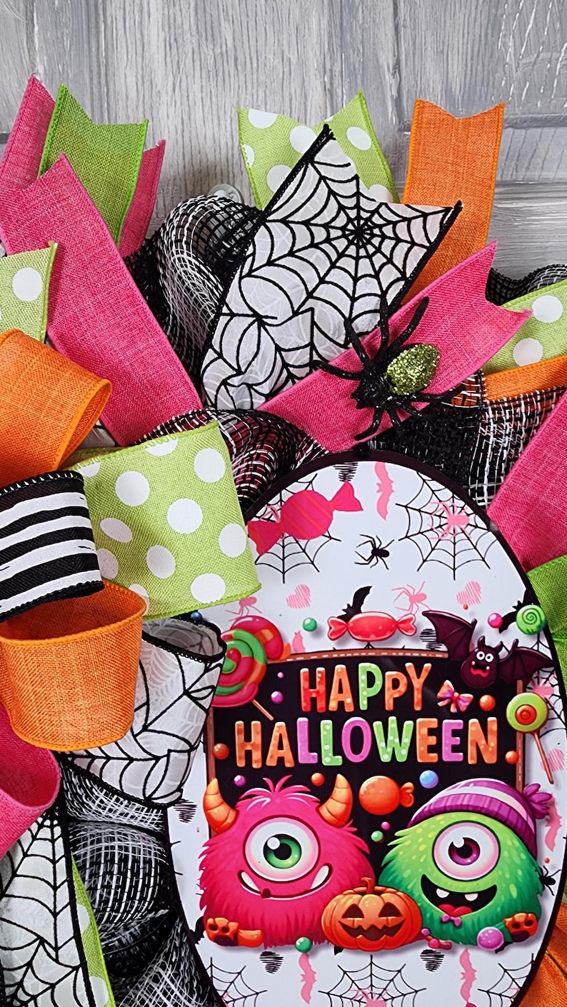 Puede incluir: Una corona de Halloween con cintas coloridas en rosa, verde y naranja. La corona presenta un letrero que dice "Happy Halloween" con monstruos de dibujos animados, murci&eacute;lagos y caramelos. Tambi&eacute;n se incluyen ara&ntilde;as y telara&ntilde;as decorativas.