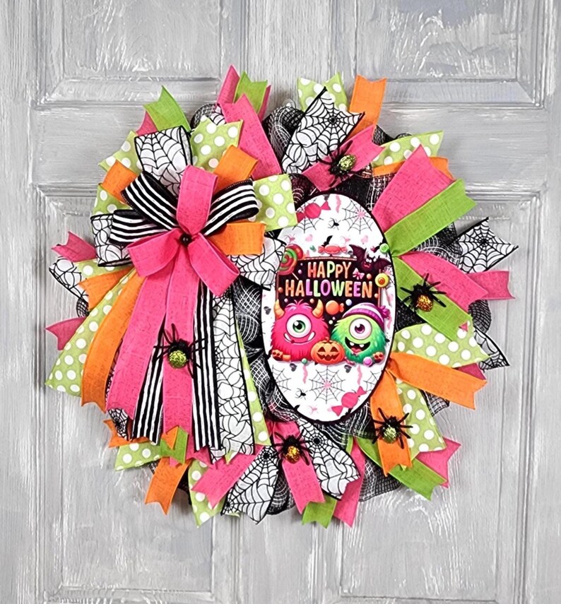 Puede incluir: Una colorida corona de Halloween con cintas rosas, naranjas, verdes y negras. La corona presenta un &oacute;valo central con las palabras "Happy Halloween" y monstruos de dibujos animados. Se adjuntan ara&ntilde;as decorativas.