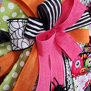 Puede incluir: Una corona de Halloween con una variedad de cintas coloridas. La corona presenta cintas rosas, naranjas, verdes y negras y blancas a rayas y lunares. Un gran lazo rosa es el punto focal, con una ara&ntilde;a negra brillante adjunta. Un gr&aacute;fico tem&aacute;tico de Halloween es visible.