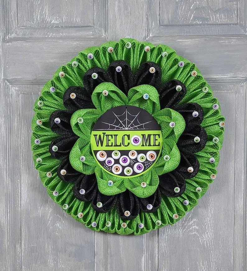 May include: A green and black Halloween wreath with a spiderweb and the word "WELCOME" on a black circle. The wreath is decorated with googly eyes.