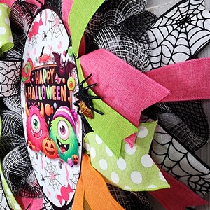 Puede incluir: Una colorida corona de Halloween con cintas rosas, verdes, naranjas y negras. La corona presenta un &oacute;valo central con las palabras "Happy Halloween" y monstruos de dibujos animados. Se adjuntan ara&ntilde;as decorativas.