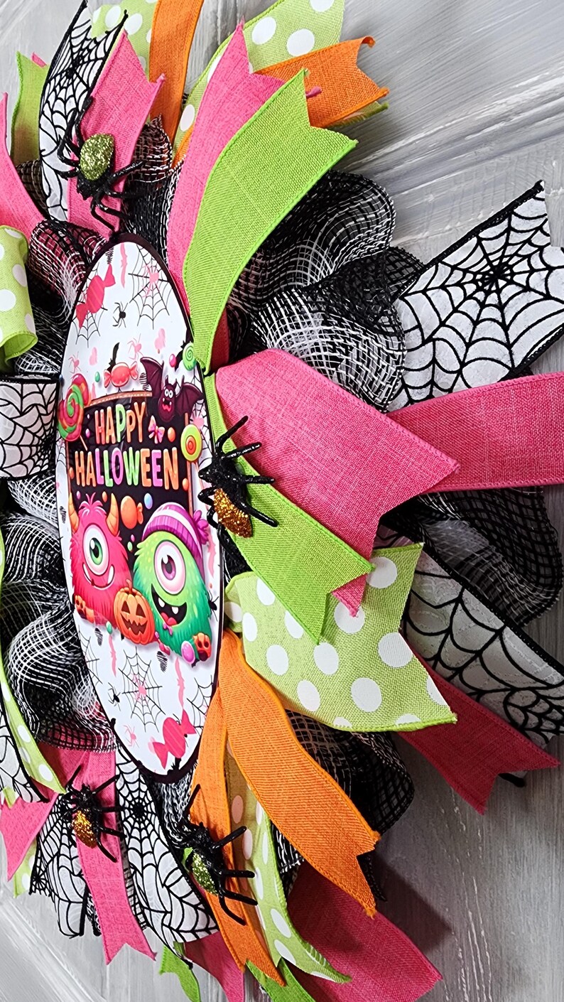 Puede incluir: Una colorida corona de Halloween con cintas rosas, verdes, naranjas y negras. La corona presenta un &oacute;valo central con las palabras "Happy Halloween" y monstruos de dibujos animados. Se adjuntan ara&ntilde;as decorativas.