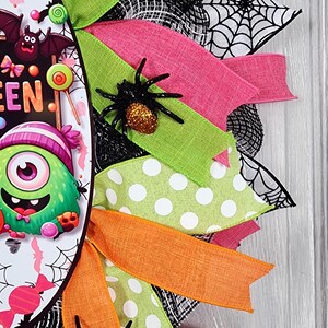 Puede incluir: Una colorida corona de Halloween con cintas rosas, verdes y naranjas. La corona presenta un dise&ntilde;o central con un monstruo de dibujos animados y la palabra "EEN". Se adjuntan ara&ntilde;as negras a las cintas y se ven detalles de telara&ntilde;as.