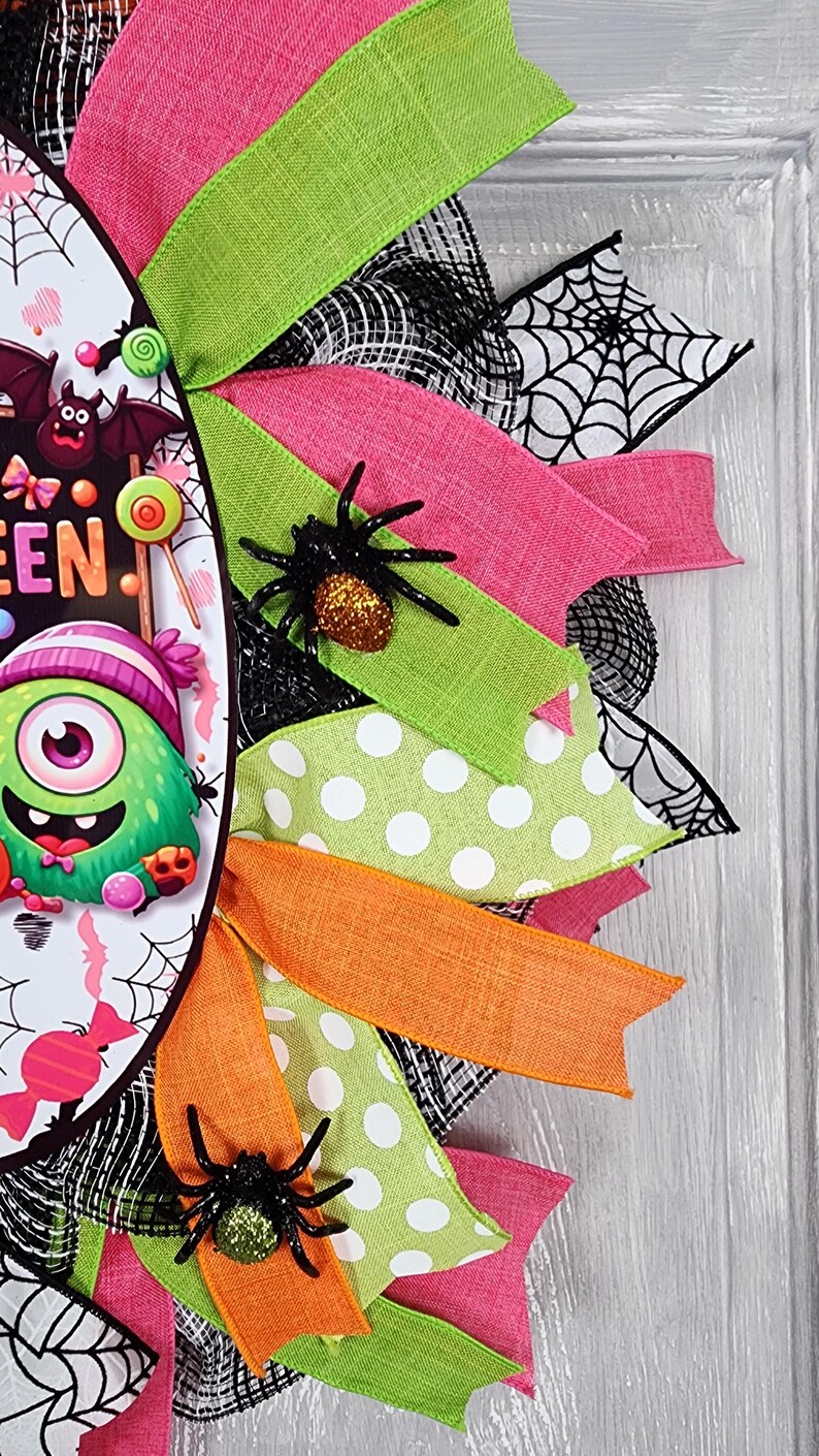 Puede incluir: Una colorida corona de Halloween con cintas rosas, verdes y naranjas. La corona presenta un dise&ntilde;o central con un monstruo de dibujos animados y la palabra "EEN". Se adjuntan ara&ntilde;as negras a las cintas y se ven detalles de telara&ntilde;as.