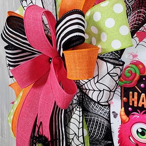 Puede incluir: Una colorida corona de Halloween con cintas rosas, naranjas, blancas y negras, y una cinta verde con lunares. La corona presenta un peque&ntilde;o cubo naranja, una ara&ntilde;a negra brillante y un letrero que dice "HA" con un monstruo.