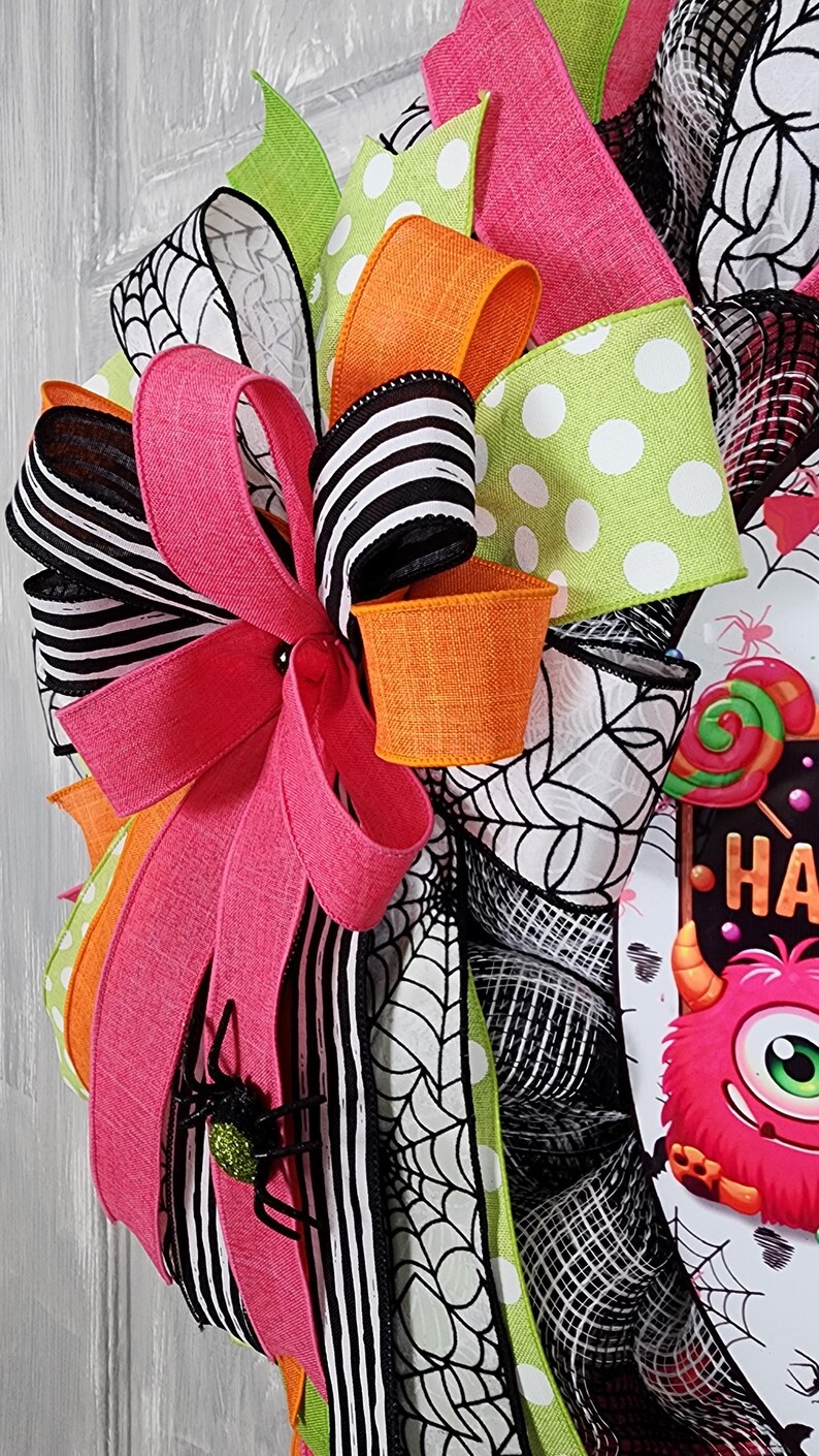 Puede incluir: Una colorida corona de Halloween con cintas rosas, naranjas, blancas y negras, y una cinta verde con lunares. La corona presenta un peque&ntilde;o cubo naranja, una ara&ntilde;a negra brillante y un letrero que dice "HA" con un monstruo.
