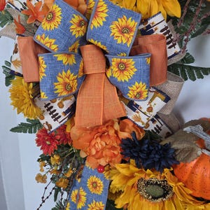 Puede incluir: Una corona decorativa con grandes girasoles amarillos, peon&iacute;as naranjas y flores azul oscuro. Un lazo central hecho de cinta azul con estampado de girasoles y cinta naranja s&oacute;lida se acent&uacute;a con cinta de arpillera con un estampado de cosecha.