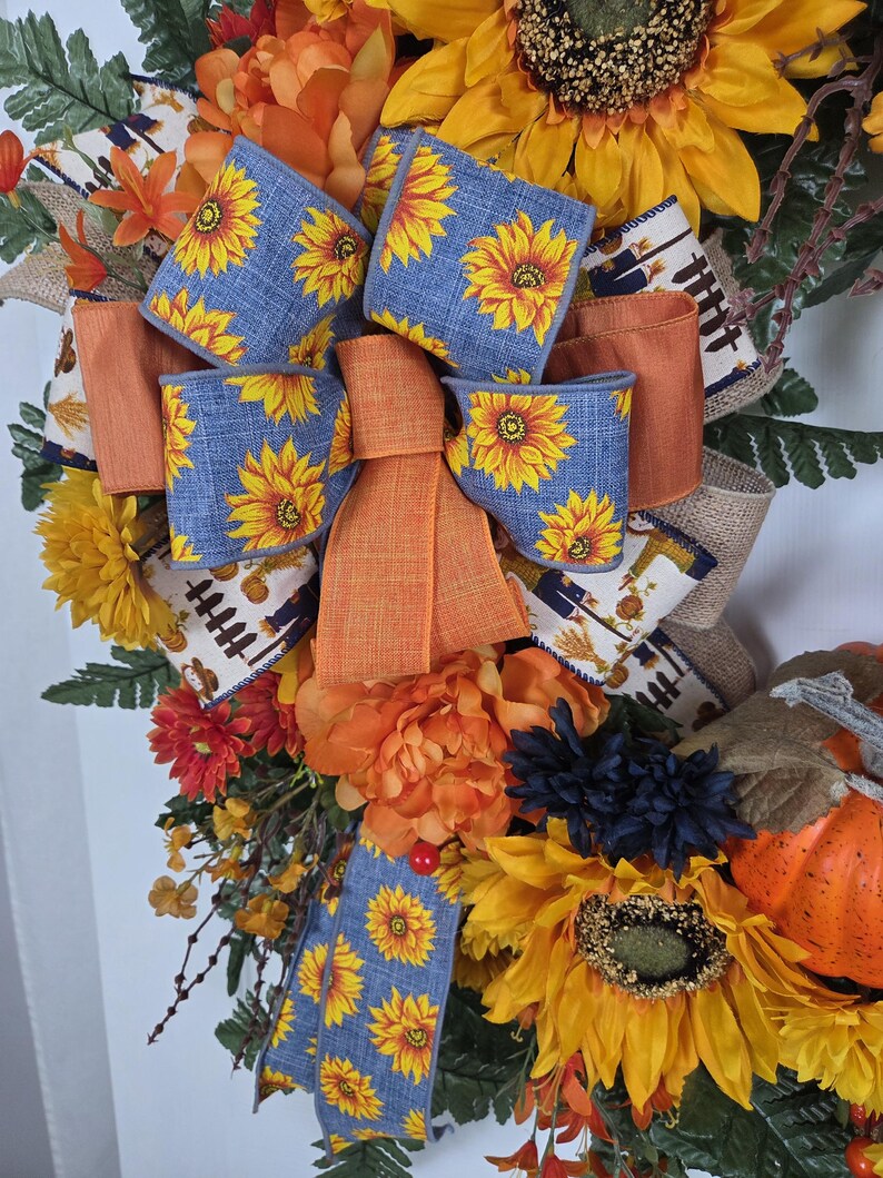 Puede incluir: Una corona decorativa con grandes girasoles amarillos, peon&iacute;as naranjas y flores azul oscuro. Un lazo central hecho de cinta azul con estampado de girasoles y cinta naranja s&oacute;lida se acent&uacute;a con cinta de arpillera con un estampado de cosecha.