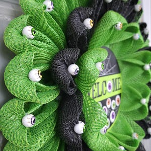 May include: A green and black Halloween wreath with a welcome sign and googly eyes.