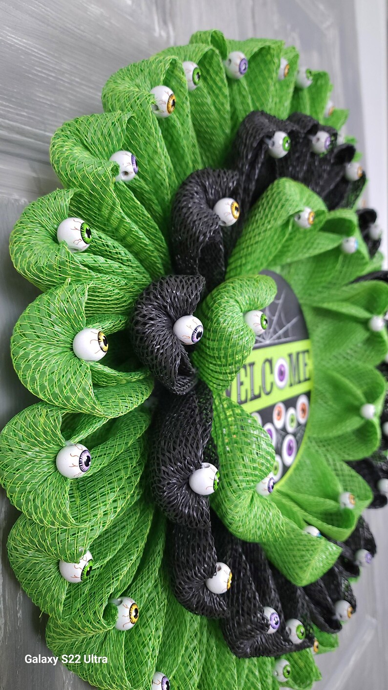 May include: A green and black Halloween wreath with a welcome sign and googly eyes.