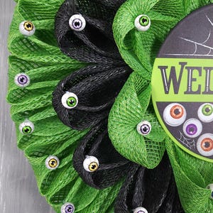 May include: A green and black mesh wreath decorated with googly eyes. The wreath has a sign that says "Welcome".