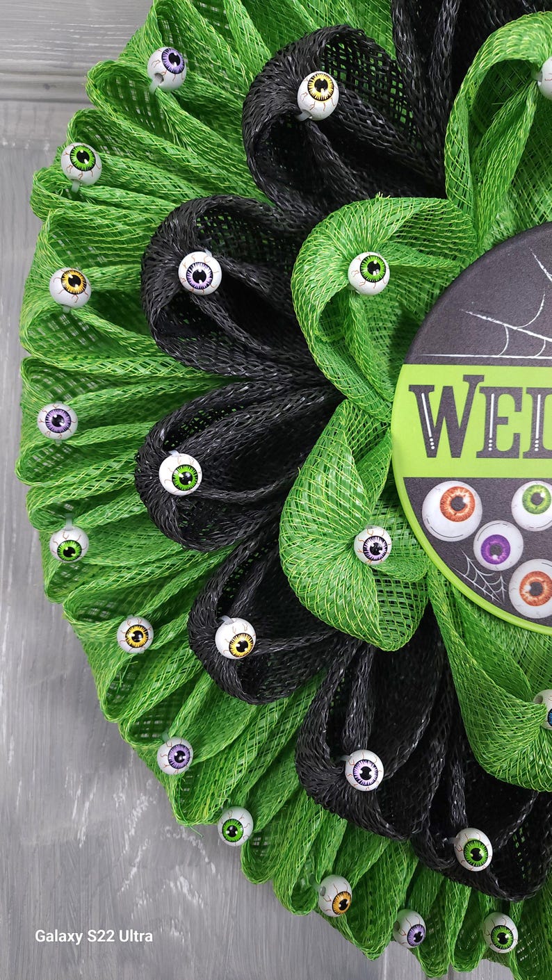 May include: A green and black mesh wreath decorated with googly eyes. The wreath has a sign that says "Welcome".
