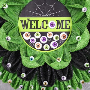 May include: A black and green Halloween wreath with a welcome sign and googly eyes. The sign says "Welcome" in white letters with a spiderweb design.