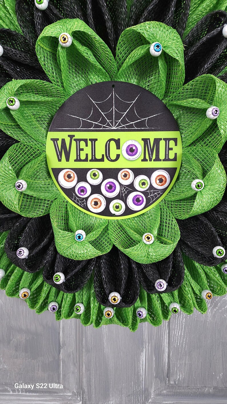 May include: A black and green Halloween wreath with a welcome sign and googly eyes. The sign says "Welcome" in white letters with a spiderweb design.