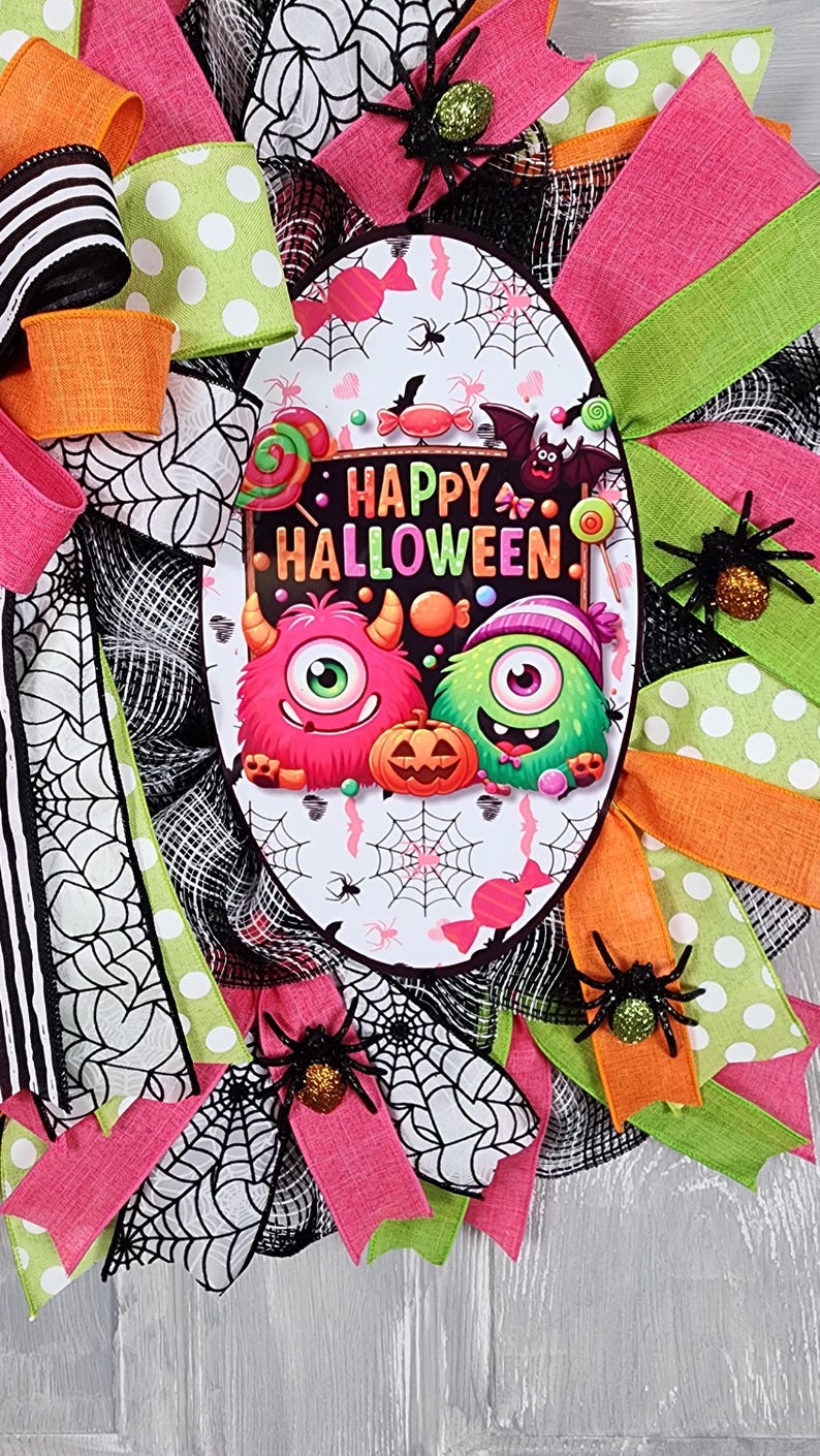 Puede incluir: Una corona de Halloween con un &oacute;valo central con monstruos de dibujos animados y el texto "HAPPY HALLOWEEN". La corona est&aacute; decorada con cintas rosas, verdes, naranjas y negras, telara&ntilde;as y ara&ntilde;as brillantes. Una decoraci&oacute;n festiva y colorida.