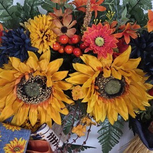 Puede incluir: Una corona decorativa con grandes girasoles amarillos, crisantemos azul oscuro, margaritas naranjas y bayas rojas. El arreglo incluye hojas de helecho verdes y est&aacute; acentuado con una cinta azul con estampado de girasoles y tela de arpillera.