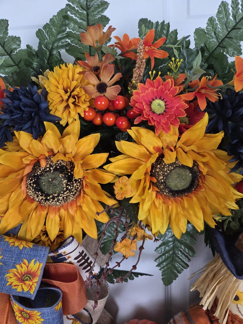 Puede incluir: Una corona decorativa con grandes girasoles amarillos, crisantemos azul oscuro, margaritas naranjas y bayas rojas. El arreglo incluye hojas de helecho verdes y est&aacute; acentuado con una cinta azul con estampado de girasoles y tela de arpillera.