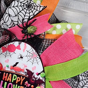 Puede incluir: Una colorida corona de Halloween con un dise&ntilde;o circular. La corona presenta cintas rosas, verdes, naranjas y blancas con lunares. Un letrero central dice "Happy Halloween" con monstruos de dibujos animados, ara&ntilde;as y caramelos.