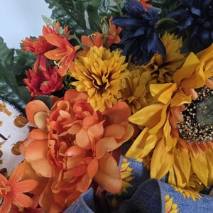Puede incluir: Un arreglo floral decorativo con peon&iacute;as naranjas, girasoles amarillos y flores azul oscuro mezcladas con follaje verde. Una cinta azul con un patr&oacute;n de girasoles amarillos est&aacute; integrada en la base, junto a un detalle de tela blanca.