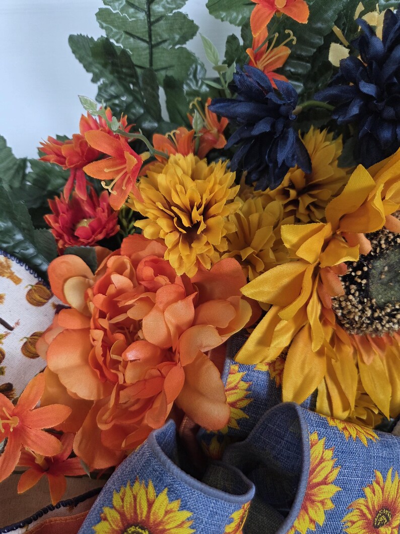 Puede incluir: Un arreglo floral decorativo con peon&iacute;as naranjas, girasoles amarillos y flores azul oscuro mezcladas con follaje verde. Una cinta azul con un patr&oacute;n de girasoles amarillos est&aacute; integrada en la base, junto a un detalle de tela blanca.