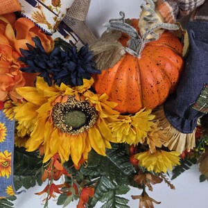Puede incluir: Una corona decorativa que presenta una calabaza naranja brillante con un tallo texturizado, grandes girasoles amarillos, flores azul oscuro y follaje verde. El arreglo incluye cintas de tela estampadas con motivos de espantap&aacute;jaros y detalles a cuadros.