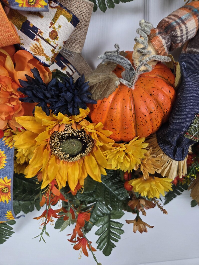 Puede incluir: Una corona decorativa que presenta una calabaza naranja brillante con un tallo texturizado, grandes girasoles amarillos, flores azul oscuro y follaje verde. El arreglo incluye cintas de tela estampadas con motivos de espantap&aacute;jaros y detalles a cuadros.