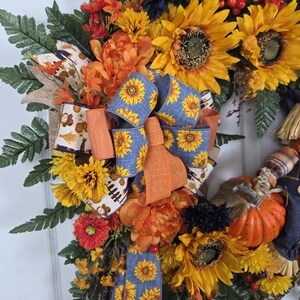 Puede incluir: Una corona decorativa con girasoles amarillos, flores naranjas y flores azul oscuro mezcladas con hojas de helecho verdes. Un lazo central hecho de cinta azul con un patr&oacute;n de girasol y acentos de cinta naranja s&oacute;lida se asienta sobre una peque&ntilde;a calabaza naranja.