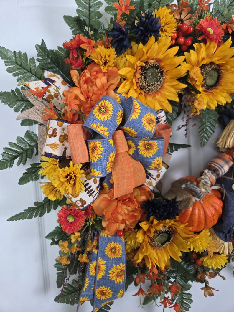 Puede incluir: Una corona decorativa con girasoles amarillos, flores naranjas y flores azul oscuro mezcladas con hojas de helecho verdes. Un lazo central hecho de cinta azul con un patr&oacute;n de girasol y acentos de cinta naranja s&oacute;lida se asienta sobre una peque&ntilde;a calabaza naranja.