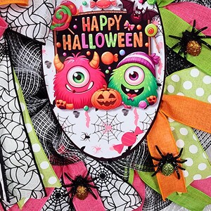 Puede incluir: Una corona de Halloween con un letrero ovalado central que dice "Happy Halloween". El letrero presenta monstruos de dibujos animados, caramelos y una calabaza. La corona est&aacute; decorada con cintas rosas, verdes y naranjas, telara&ntilde;as y ara&ntilde;as brillantes.