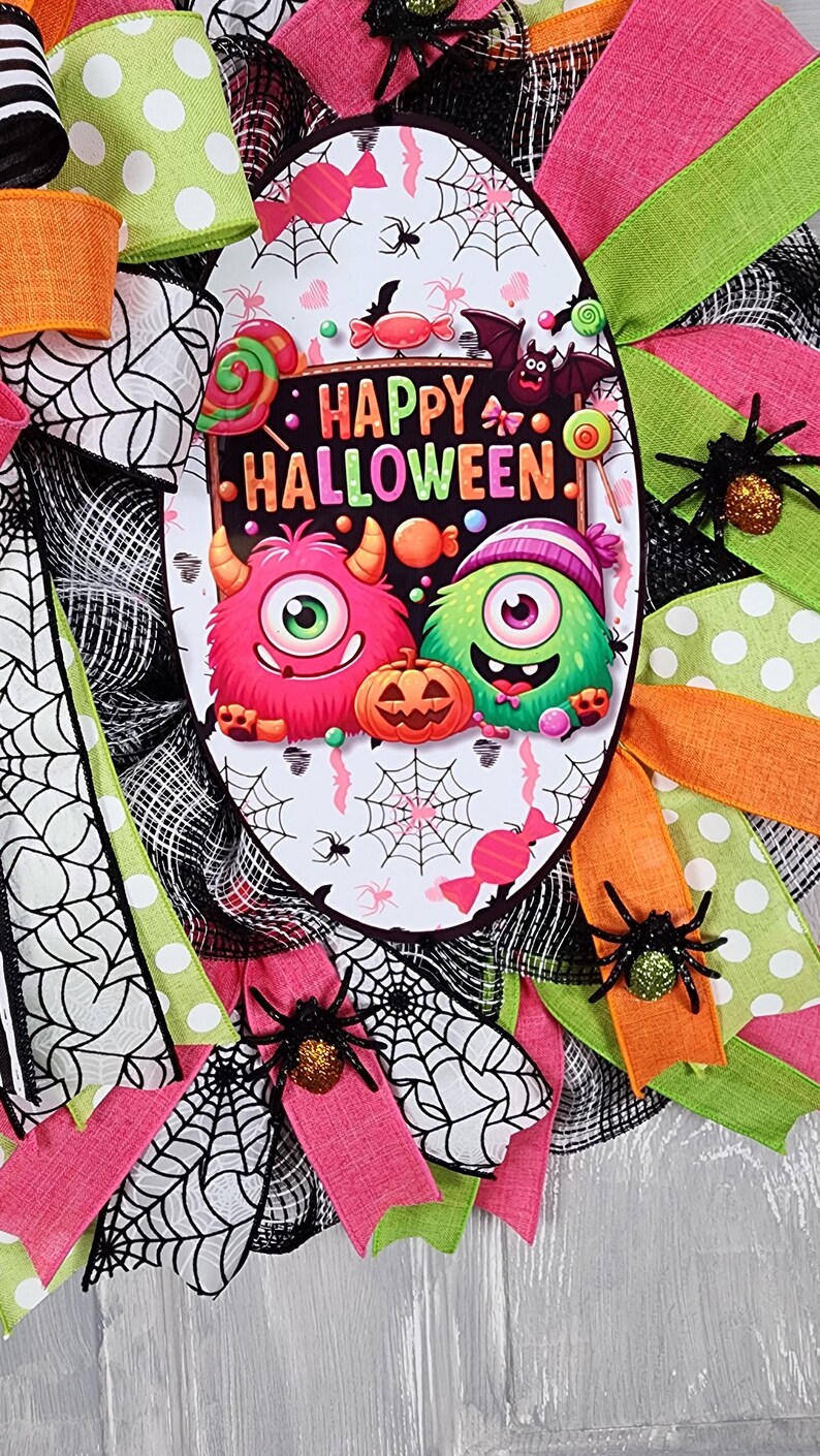 Puede incluir: Una corona de Halloween con un letrero ovalado central que dice "Happy Halloween". El letrero presenta monstruos de dibujos animados, caramelos y una calabaza. La corona est&aacute; decorada con cintas rosas, verdes y naranjas, telara&ntilde;as y ara&ntilde;as brillantes.