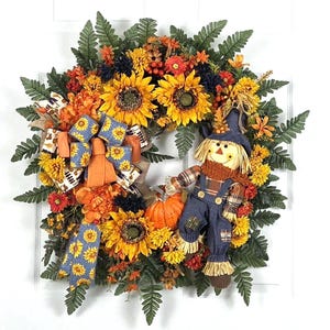 Puede incluir: Una corona decorativa de oto&ntilde;o con girasoles amarillos, flores naranjas, hojas de helecho verdes y una peque&ntilde;a calabaza naranja. Incluye una figura de espantap&aacute;jaros de tela con sombrero y peto azul, decorada con un lazo azul estampado.